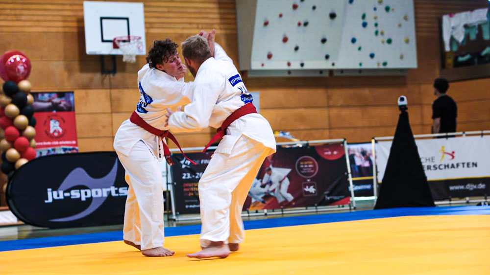 Steffen Bienias wehrt einen Schlagangriff von Tjark Rose bei der Deutschen Meisterschaft Ju-Jutsu 2026 auf der gelben Wettkampfmatte ab, beide im weißen Gi mit rotem Gürtel