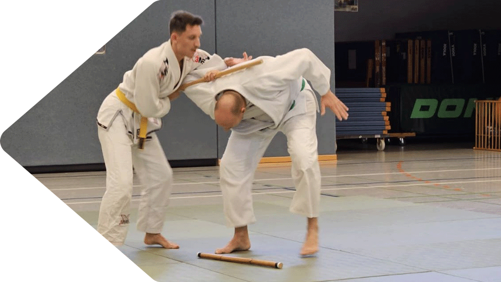 Julian und Roman bei einer Hanbo-Jutsu-Prüfung im weißen Gi. Julian mit gelbem und Roman mit grünem Gürtel demonstrieren eine kontrollierte Hanbo-Technik, bei der Romans Arm mit dem Hanbo fixiert wird. Hanbo-Jutsu-Training in Süpplingen.