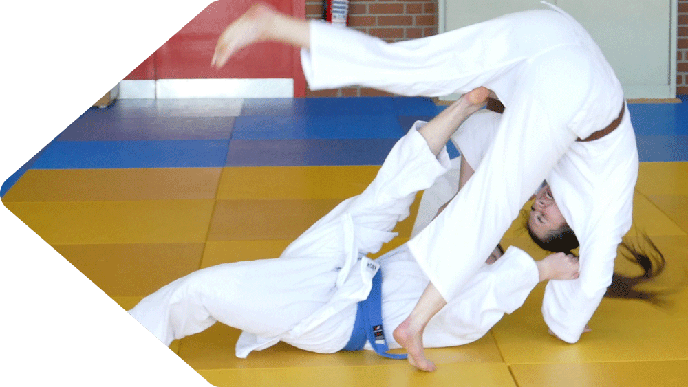 Steffen im weißen Gi mit blauem Gürtel, auf gelben Judo-Matten, wirft Finja mit einer kontrollierten Judo-Technik über sich. Die beiden trainieren Judo in Süpplingen.